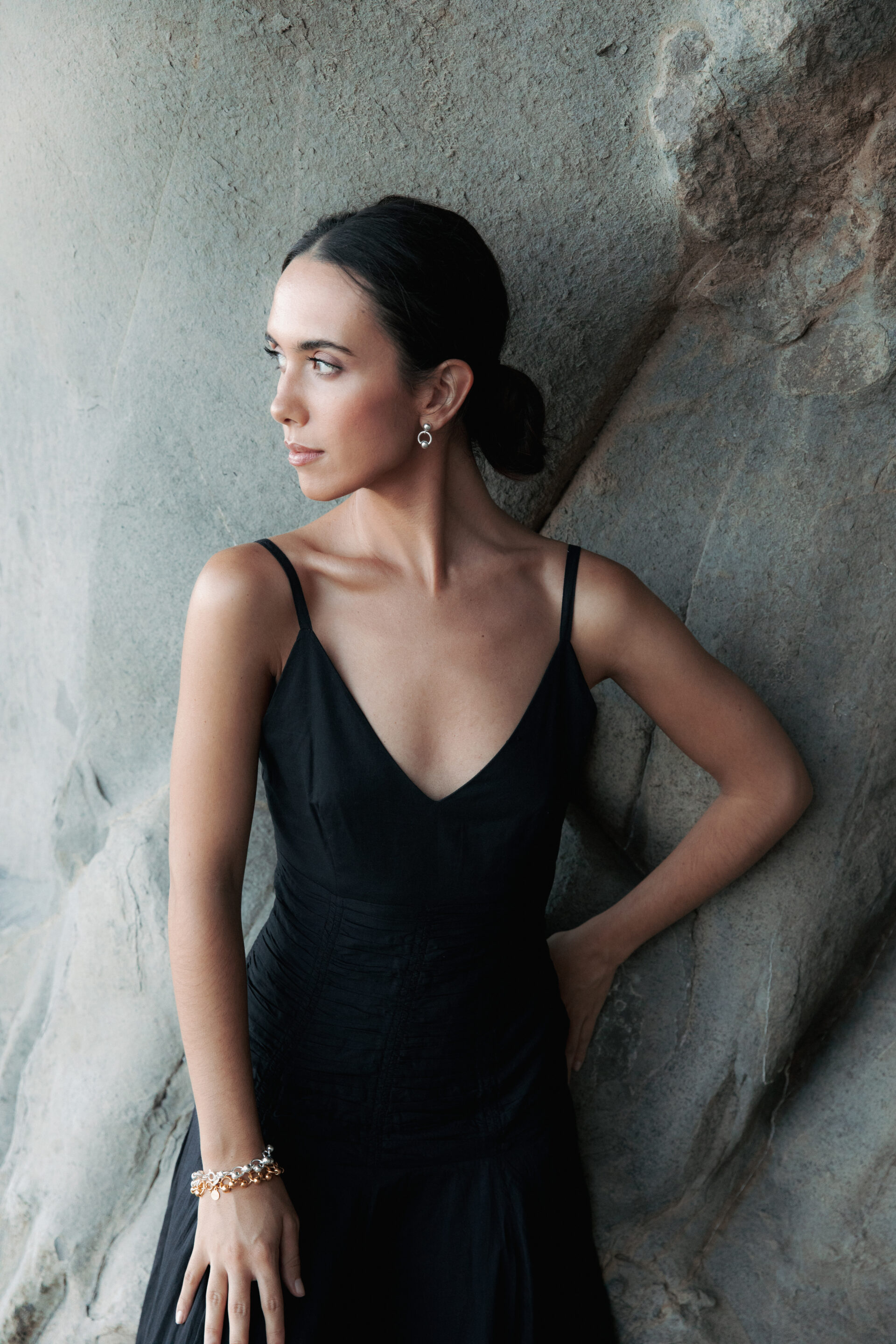 Chica con un vestido negro posando delante de un muro de rocas con la cabeza a un lado mientras fotógrafo le toma una foto